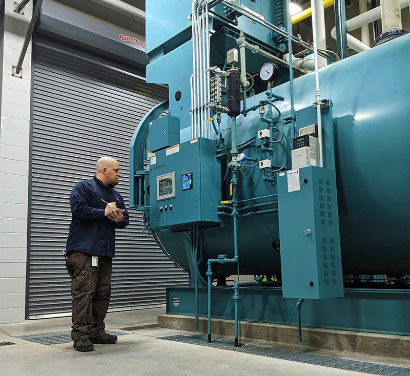 A bald man standing in front of an industrial boiler taking notes during equipment rounds at an Oatly plant.