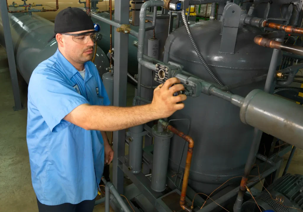 A technician wearing a blue uniform and safety glasses turns a handle on refrigerant equipment indicating ongoing maintenance or inspection