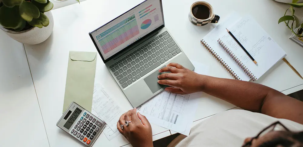 Overhead view of a woman sitting at her desk working on her budget with a calculator coffee laptop, and scattered documents in front of her