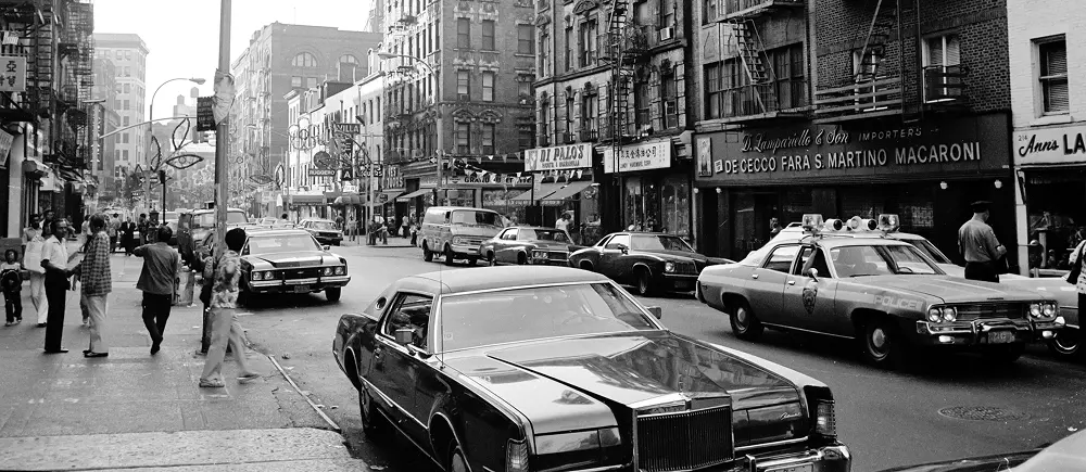 Black-and-white photo of a busy city street with pedestrians, vintage cars, a police car, and storefronts with signs for businesses and imports