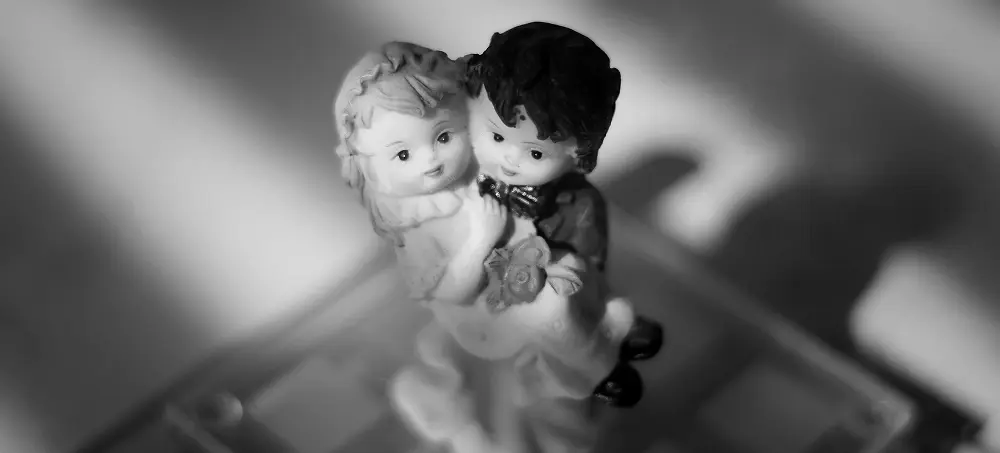Black and white photo of a ceramic figurine showing a boy and girl close together, with the boy holding flowers and leaning toward the girl, both with gentle expressions