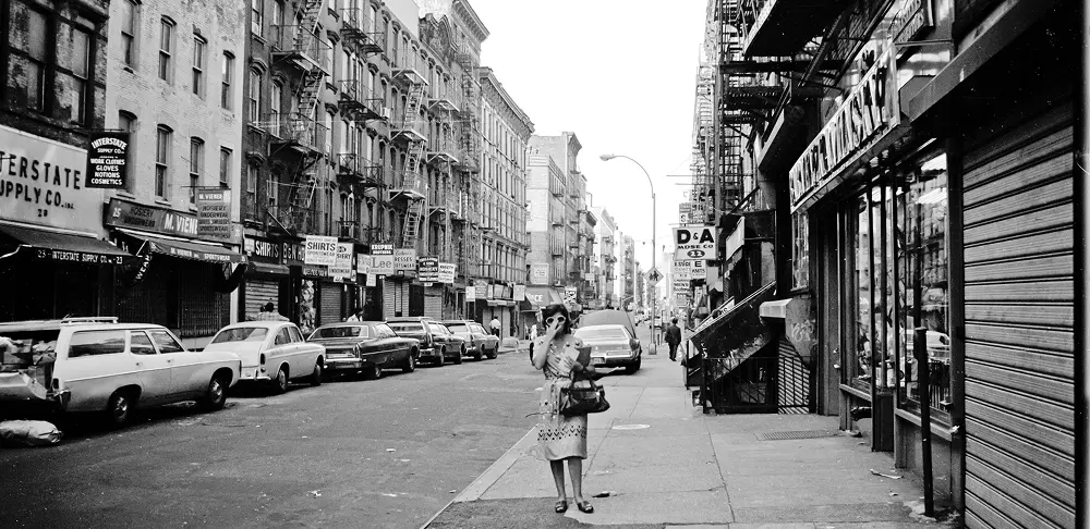 Black-and-white photo of a mid-20th century city street with parked cars, fire escape-lined apartment buildings, storefronts, and a woman standing on the sidewalk carrying bags