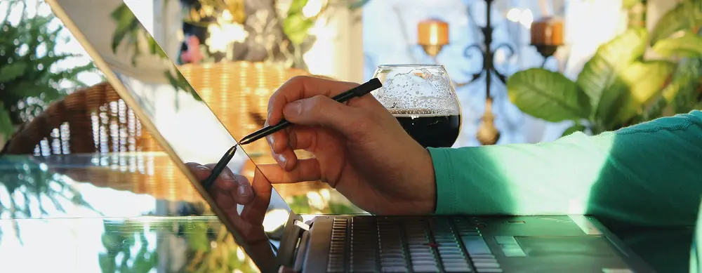 Person using a stylus on a laptop screen with a glass of dark beverage in the background