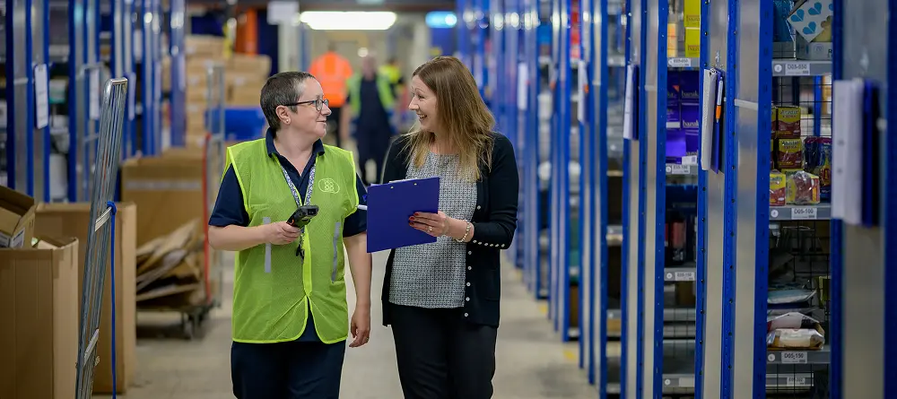 Two women in a warehouse discussing inventory, one holding a notepad, the other holding a tagging device.