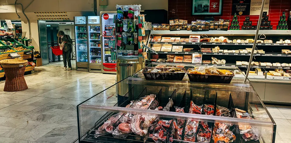 A supermarket interior with packaged meats, deli items, and drinks, with a shopper holding a red basket in the background