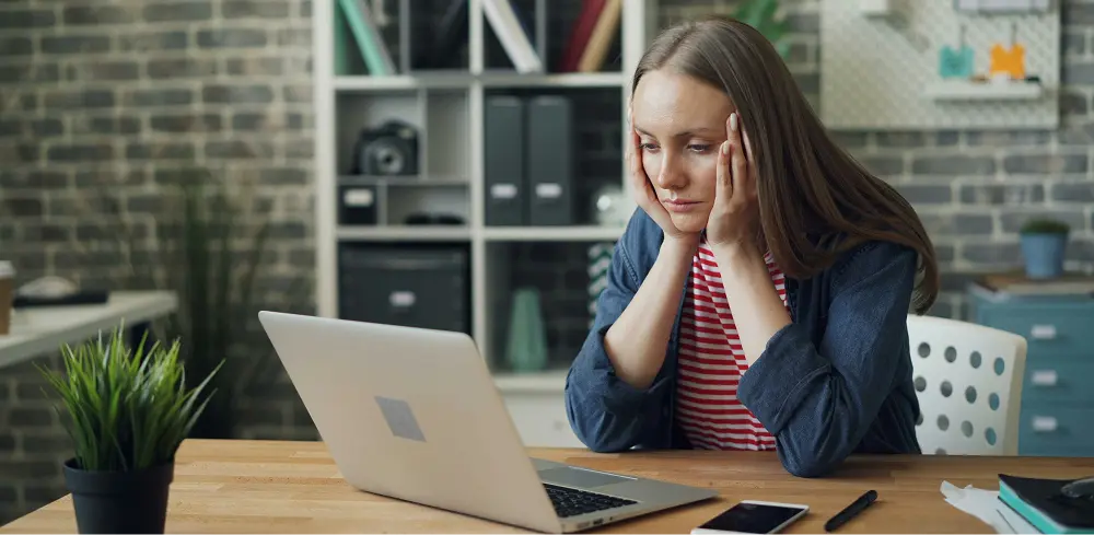 A young woman visibly stressed in her office, sitting at a desk with a laptop in front of her