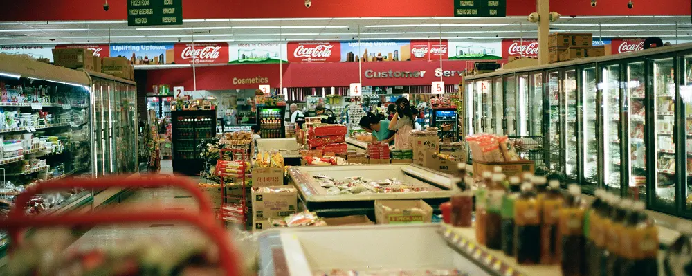 Interior of a supermarket with aisles of refrigerated display cases, food products, and shoppers at the customer service counter