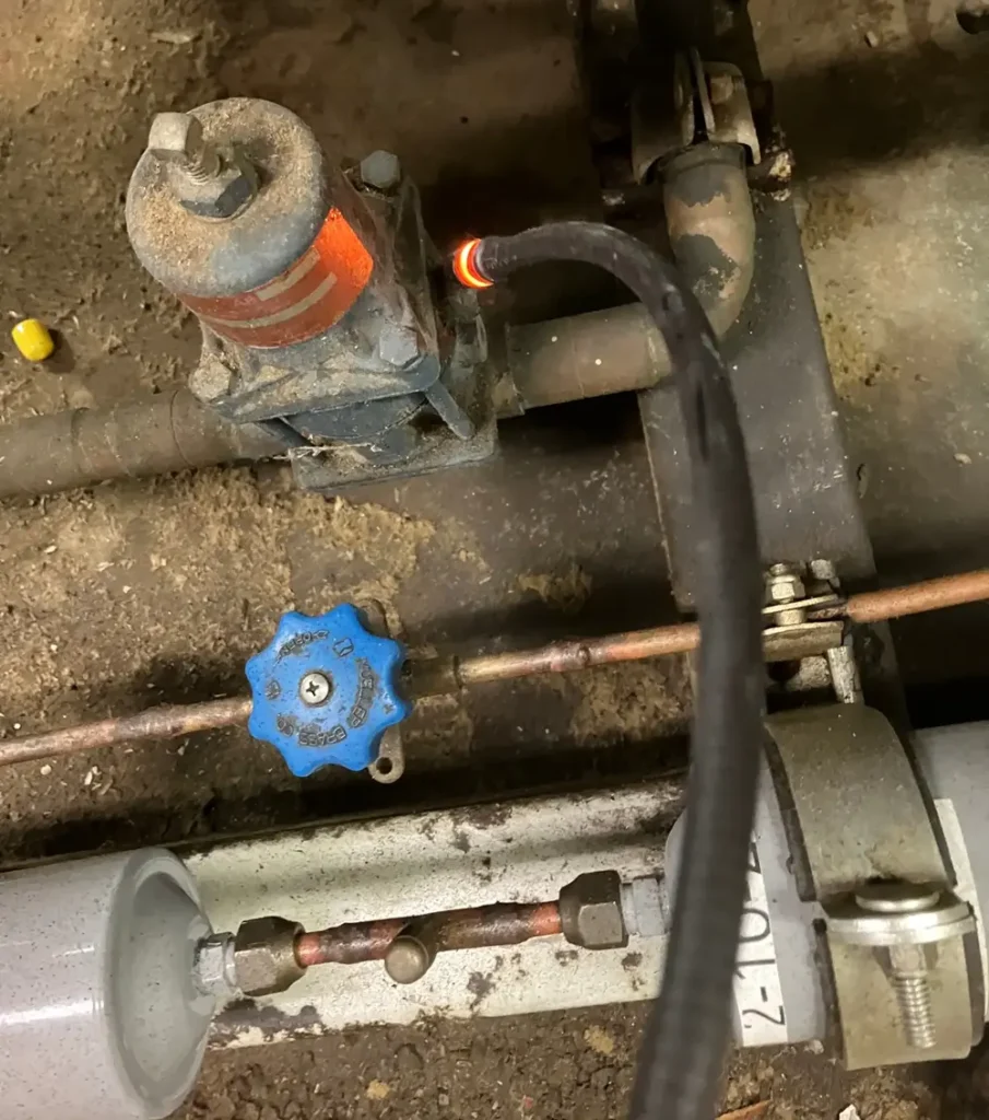 Close-up of a handheld refrigerant leak detector probe touching a corroded valve assembly on a dusty refrigeration line, with the probe tip glowing red near a blue service knob and copper tubing