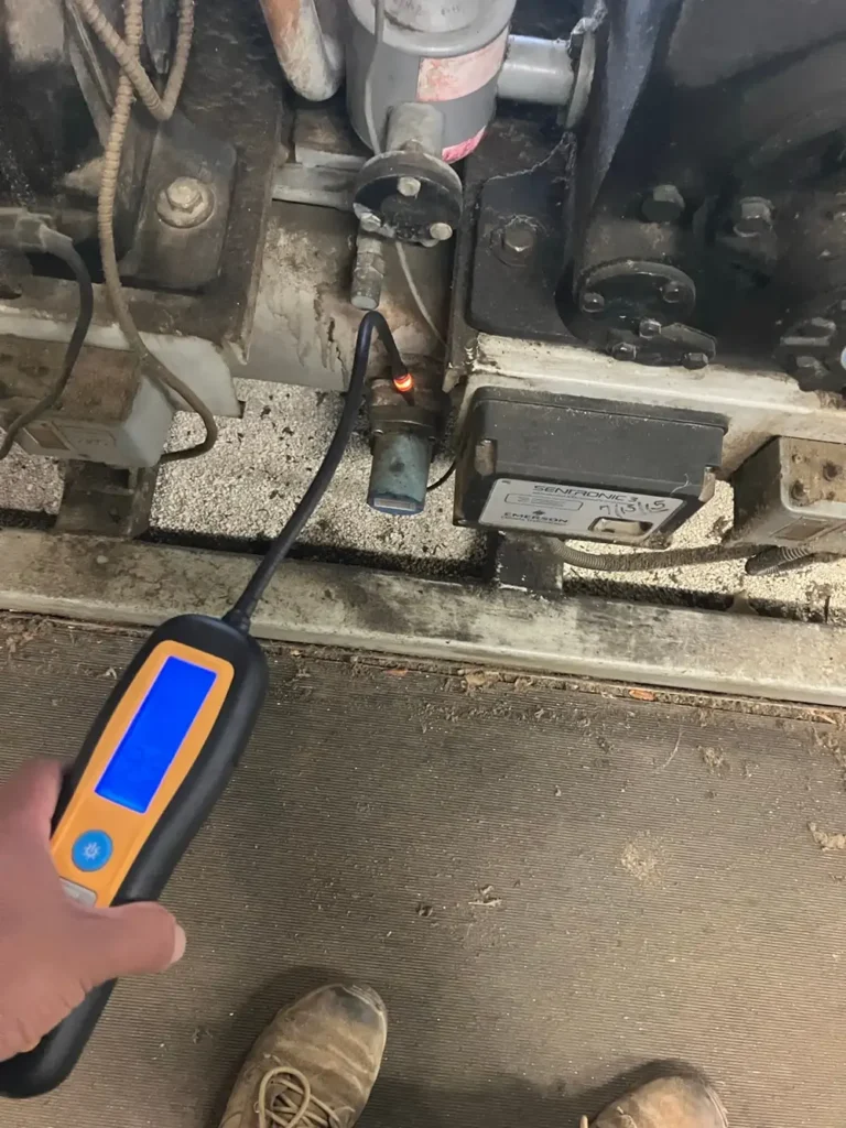 Technician holding a digital refrigerant leak detector with a blue backlit screen while the probe tests a sensor_valve on an aging compressor unit; industrial floor and worn work boots visible