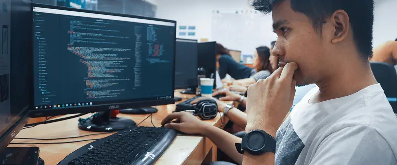 A concentrated student sits at a computer, surrounded by peers in a laptop-filled classroom