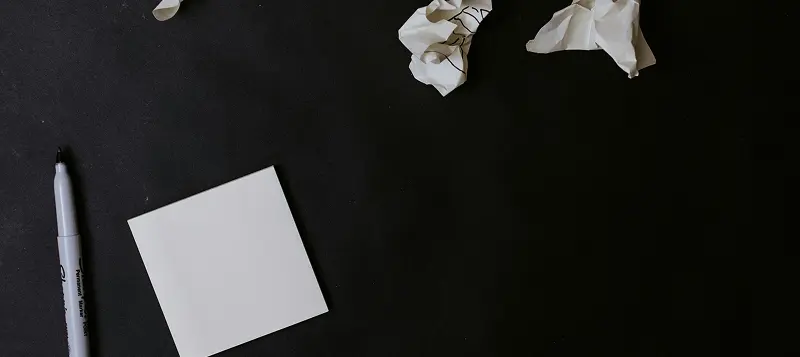 A black Sharpie marker alongside a neat stack of white sticky notes and several crumpled post-its on a flat surface