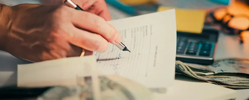 Close-up of a hand writing on a paper invoice, surrounded by scattered dollar bills and a calculator on a desk