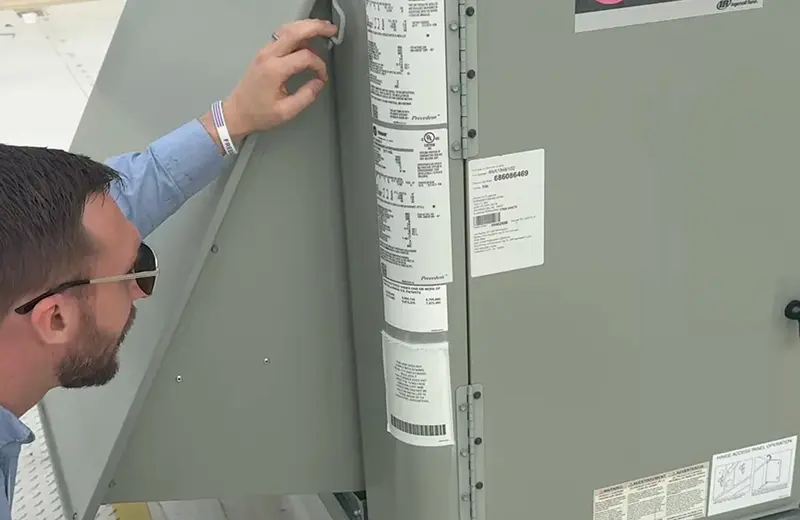 A facility manager wearing sunglasses inspects a grey industrial HVAC unit. The focus is on the unit's technical data plates and stickers, which contain barcodes and manufacturing specifications