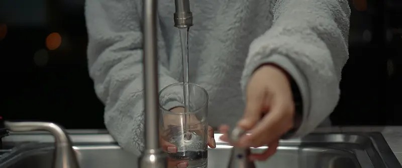 A person filling a clear glass with water from a kitchen faucet