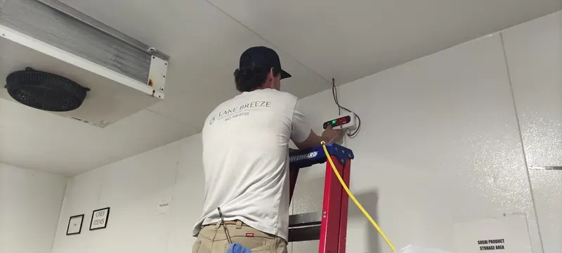 An HVAC technician stands on a red ladder inside a walk-in cooler to install an AKO leak detector on the wall The technician, wearing a Lake Breeze branded shirt, adjusts the digital display unit near the ceiling.