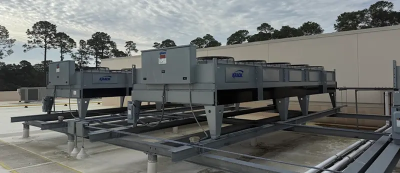 Several large, grey Krack-branded industrial condenser units mounted on steel supports on a flat commercial rooftop under a cloudy sky.