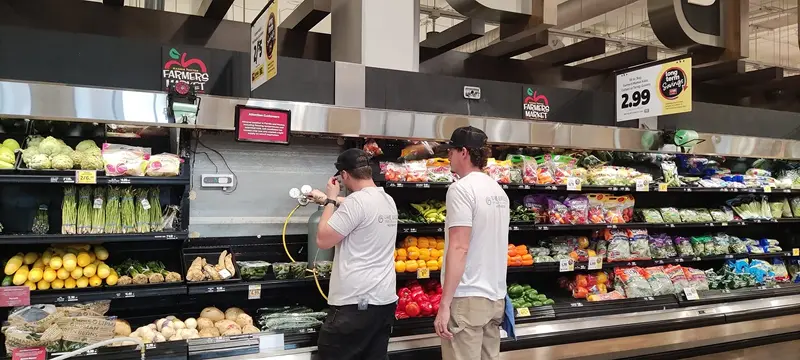 Two HVAC technicians in a grocery store work behind a produce display to install an AKO leak detector - - One technician adjusts a manifold gauge set connected by a yellow hose to the refrigerated shelving unit