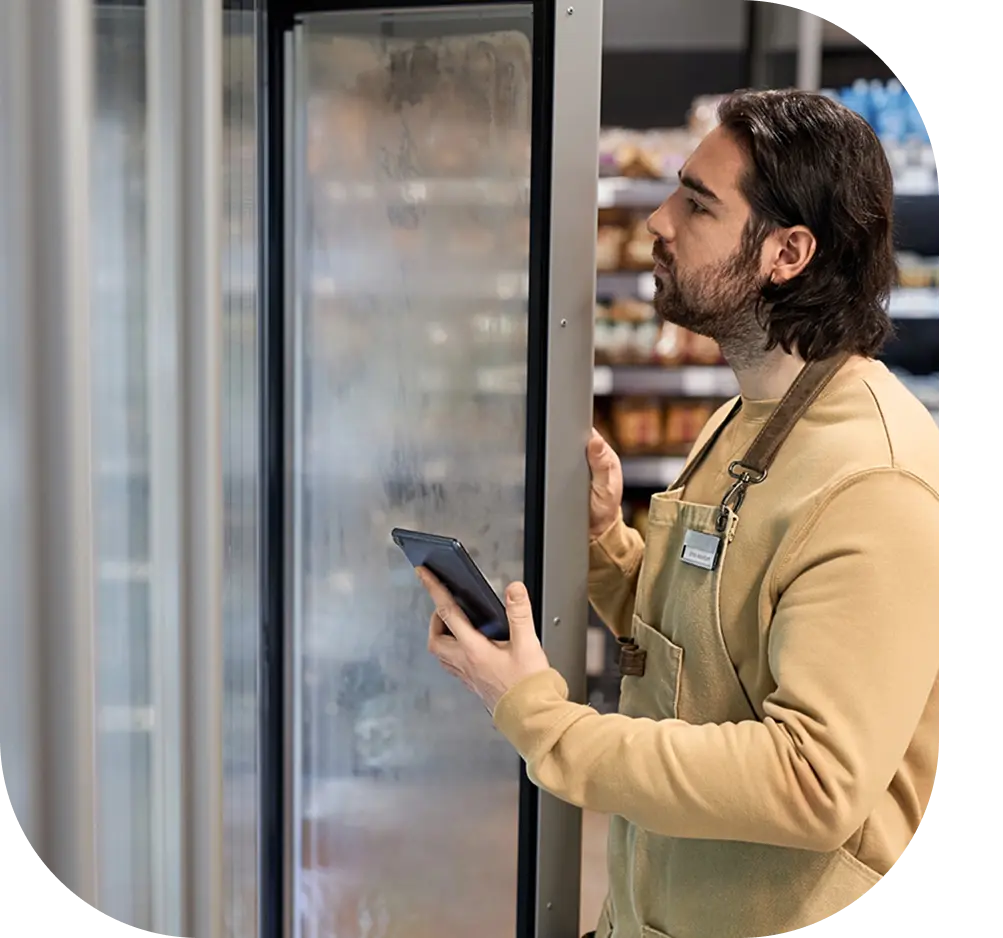 A young male worker in a supermarket is opening a refrigerant while holding a tablet in his hand wearing a beige uniform and focused on the task