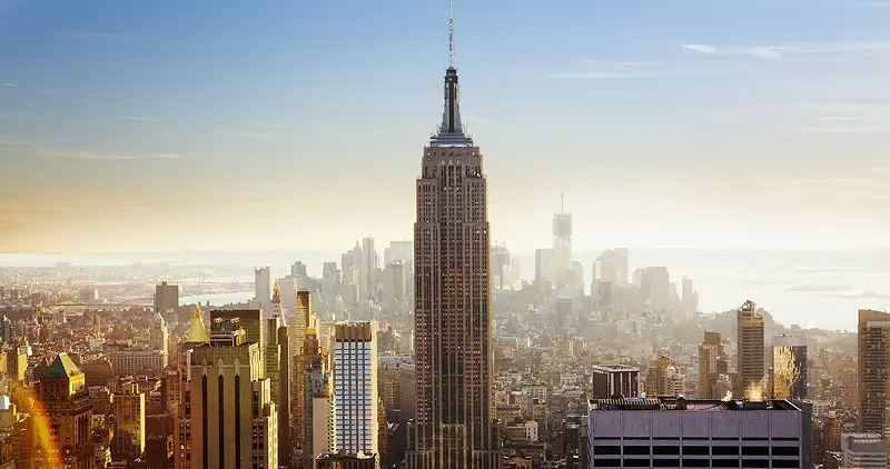 Manhattan skyscrapers catch the low golden light of sunset - their glass and stone facades glowing orange against a softening sky